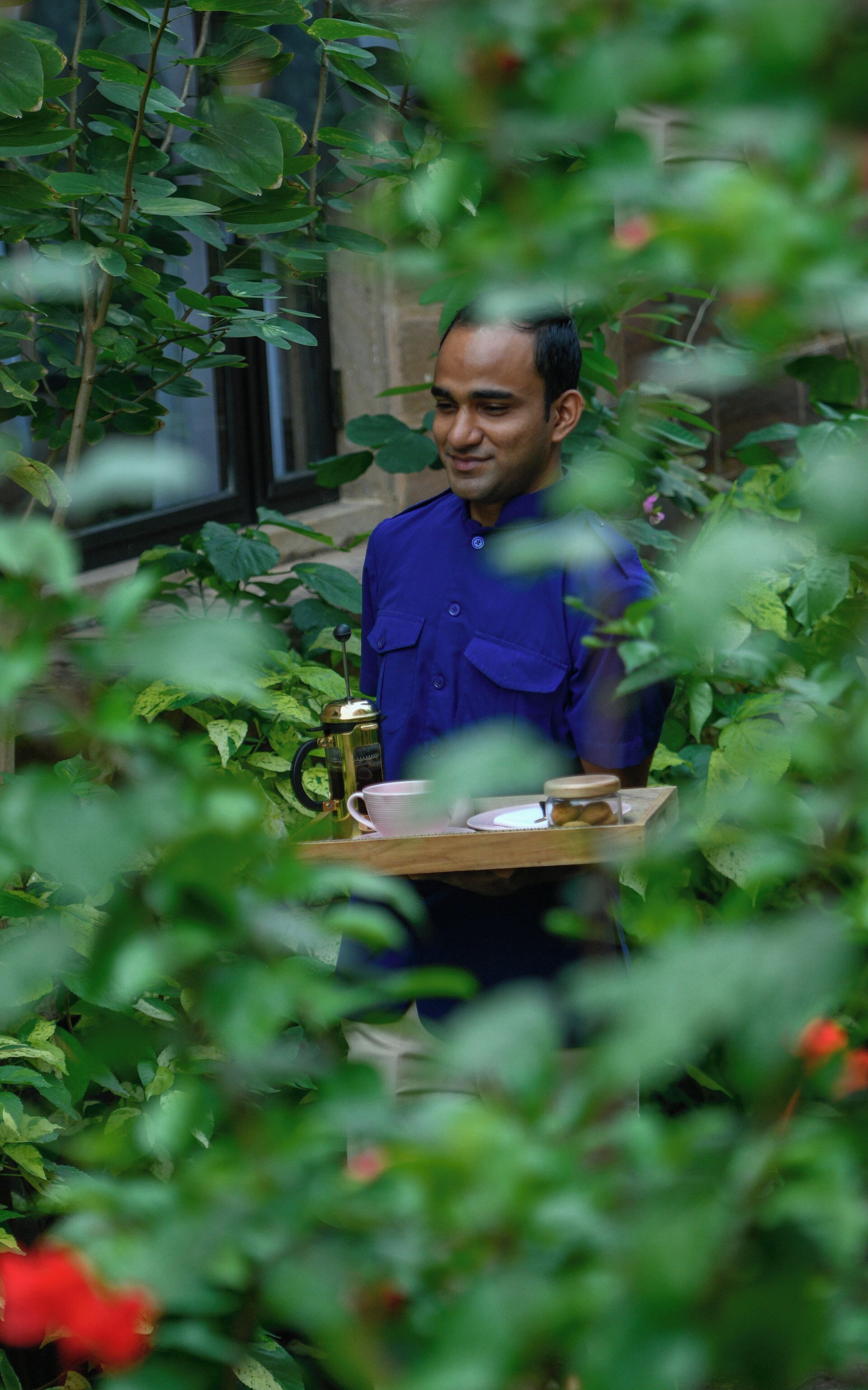 A person carrying a tray with a coffee pot, cup, and saucer, standing in a lush green garden with a building in the background.