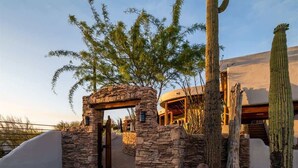 A stone archway leading to a building with a curved roof, surrounded by cacti and desert vegetation.