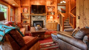A cozy log cabin interior with a fireplace, leather sofas, a television, and a staircase.