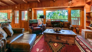 A cozy cabin interior with wooden walls, a leather sofa, a wooden coffee table, and large windows offering a view of the outdoors.