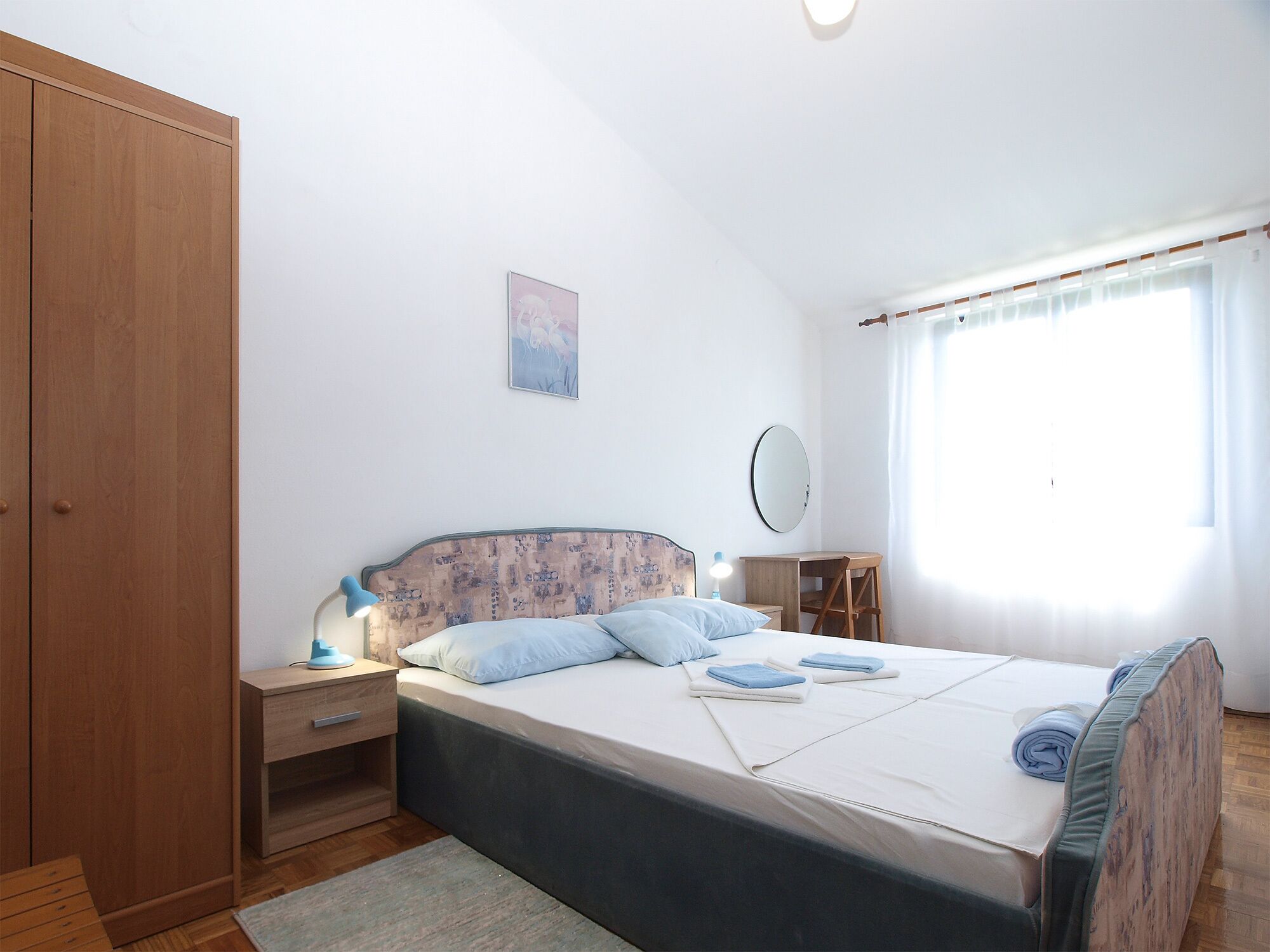 A bedroom with a bed, bedside table, nightstand, wardrobe, and a window with curtains.