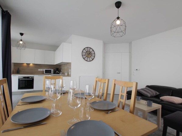 Un espace repas moderne avec une table en bois dressée pour six personnes, entourée de chaises en bois. On aperçoit une cuisine en arrière-plan, avec des armoires blanches et un four à micro-ondes.