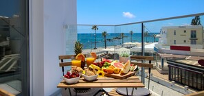 Balcón con desayunos, frutas, pan y jugo, con vista a una playa y al océano.