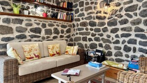 Un salon chaleureux avec un mur en pierre, un canapé beige agrémenté de coussins à motifs, un fauteuil en osier, une table blanche avec des magazines et une boîte de mouchoirs, et une bibliothèque remplie de livres.