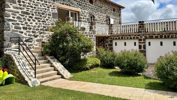Un bâtiment en pierre avec un escalier menant à une porte, le tout entouré de pelouse et d’arbustes bien entretenus.