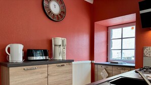Une cuisine équipée d’une machine à café, d’une bouilloire et d’un grille-pain sur le plan de travail, d’une horloge au mur et d’une fenêtre donnant sur un paysage agréable.