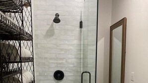 Ducha con mampara de vidrio, pared con azulejos blancos, toallero de metal y espejo con marco de madera.