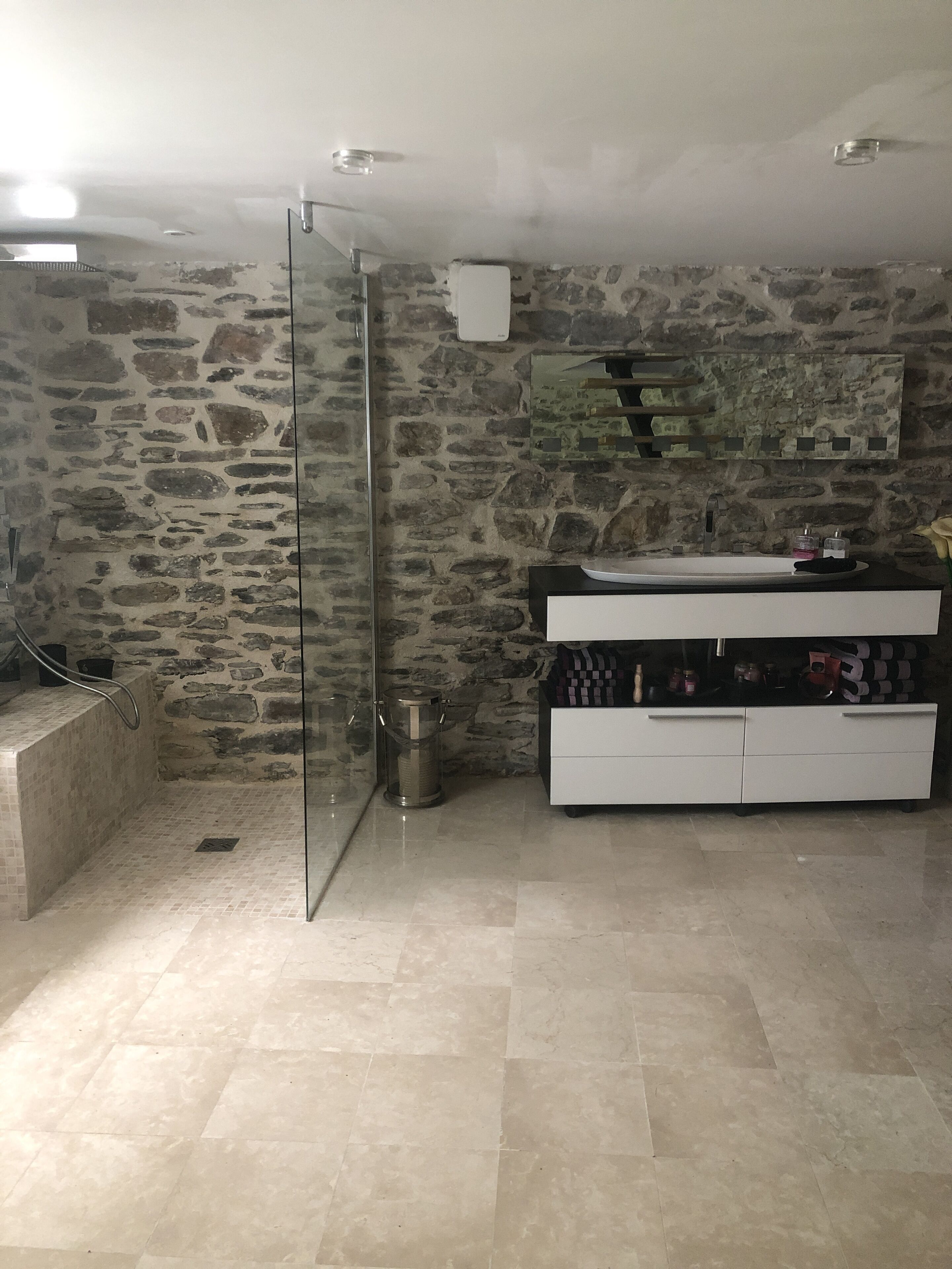 Une salle de bain avec un mur en pierre, une douche en verre, un meuble-lavabo blanc et un sol carrelé.