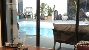 A modern living room with a wooden coffee table, a black sofa, and a glass door leading to a pool area with outdoor furniture and plants. - Villa sur la Route des Châteaux Cathares (Miraval-Cabardes)