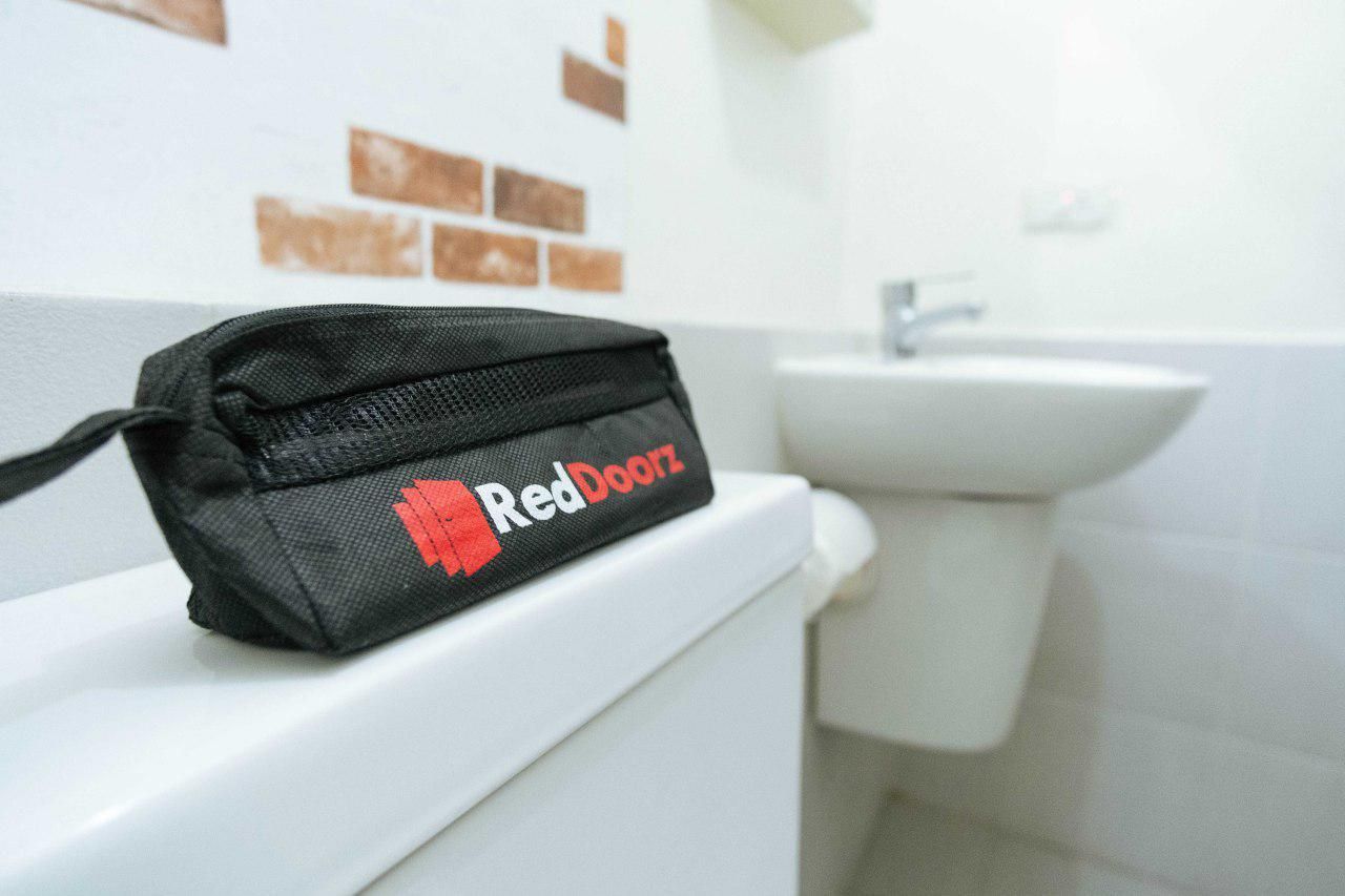 Una bolsa negra con el logotipo de "RedDoorz" sobre un inodoro blanco en un baño con lavabo y azulejos marrones en la pared.