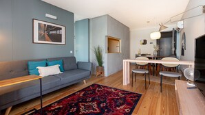A modern living room with a grey sofa, a dining table with chairs, a red patterned rug, and a large framed picture on the wall.