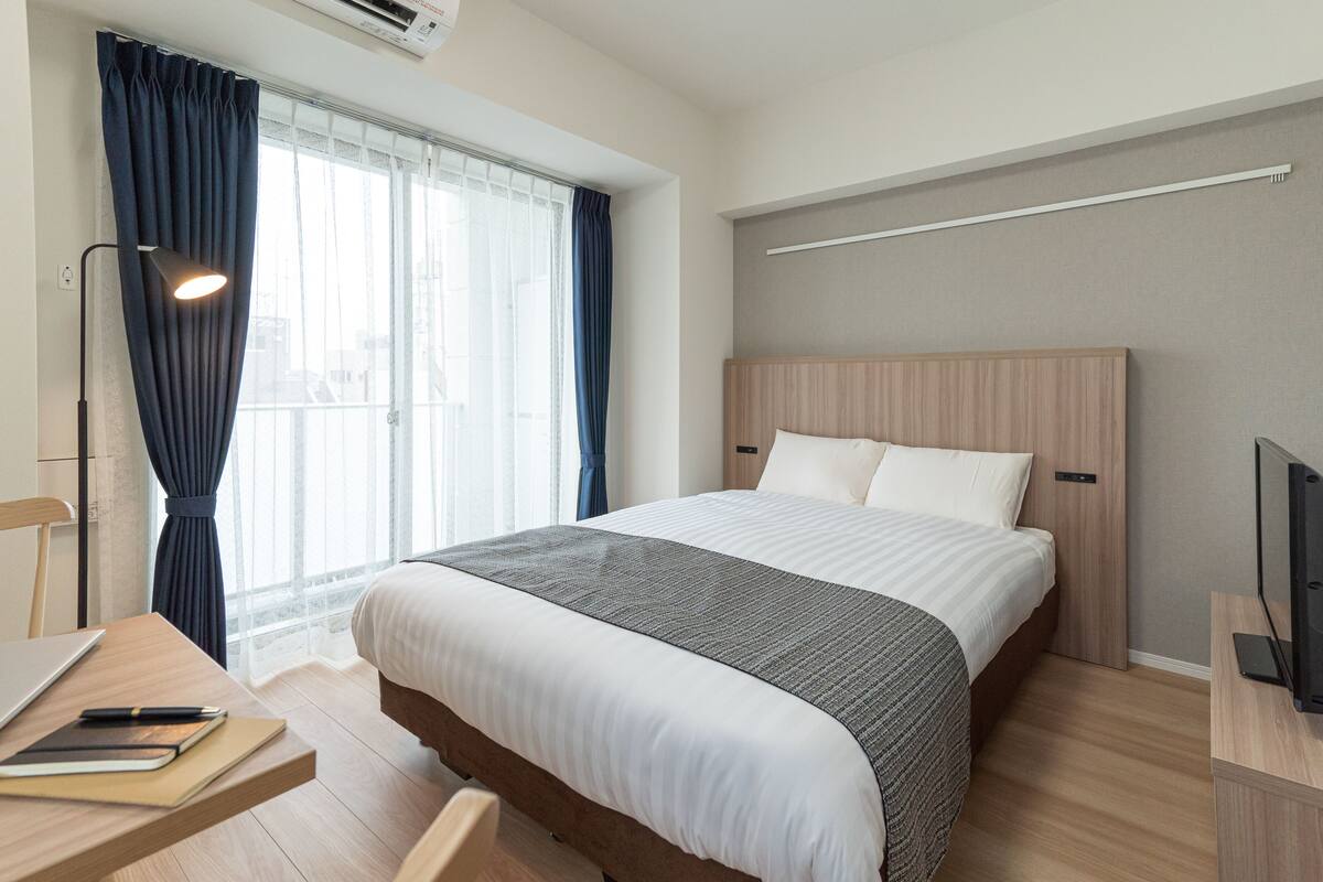 Una habitación de hotel con cama, escritorio, computadora portátil, silla, televisor y ventana con cortinas.