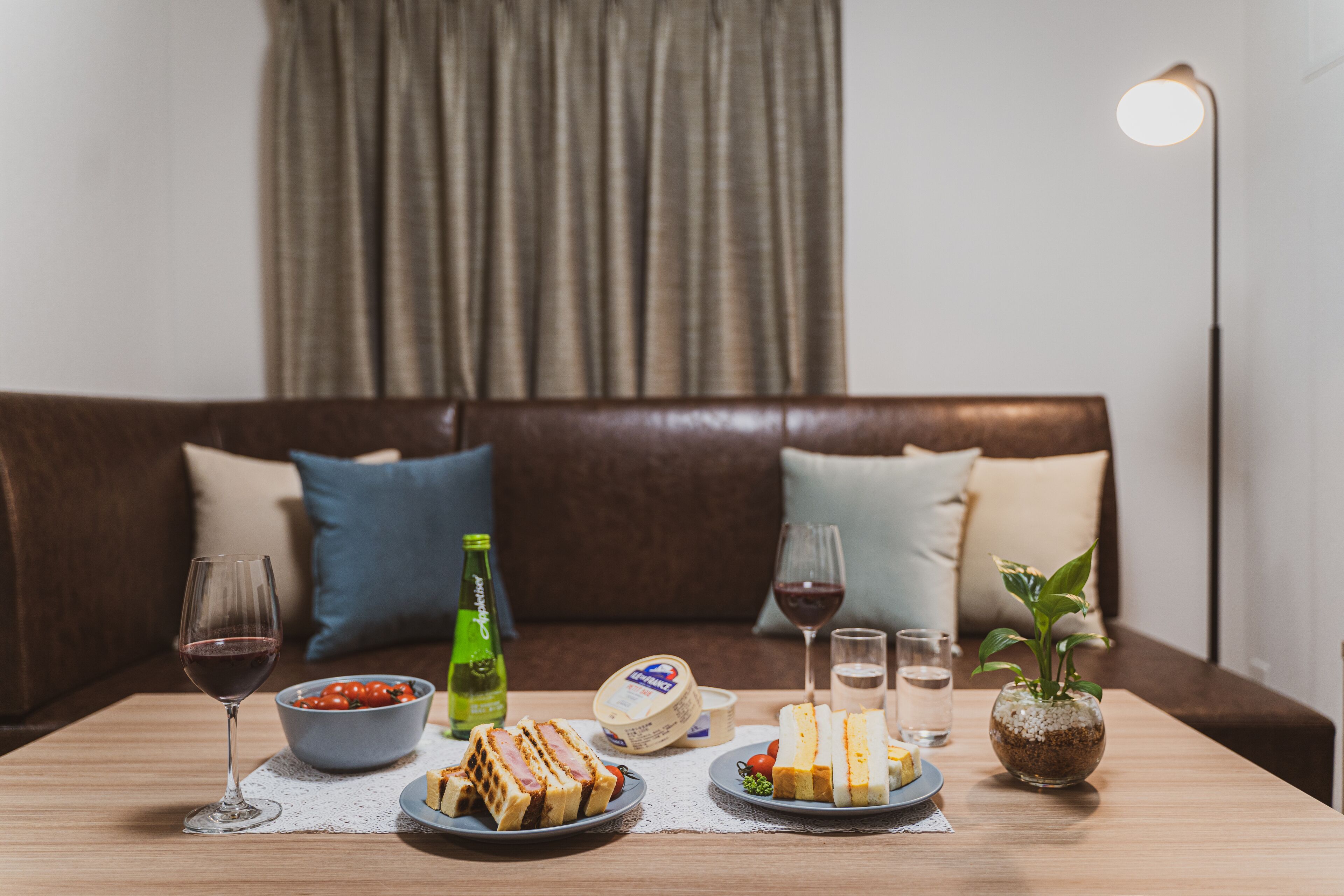 Una mesa lista con comida y bebidas en una habitación acogedora, con un sofá de cuero marrón y una lámpara.