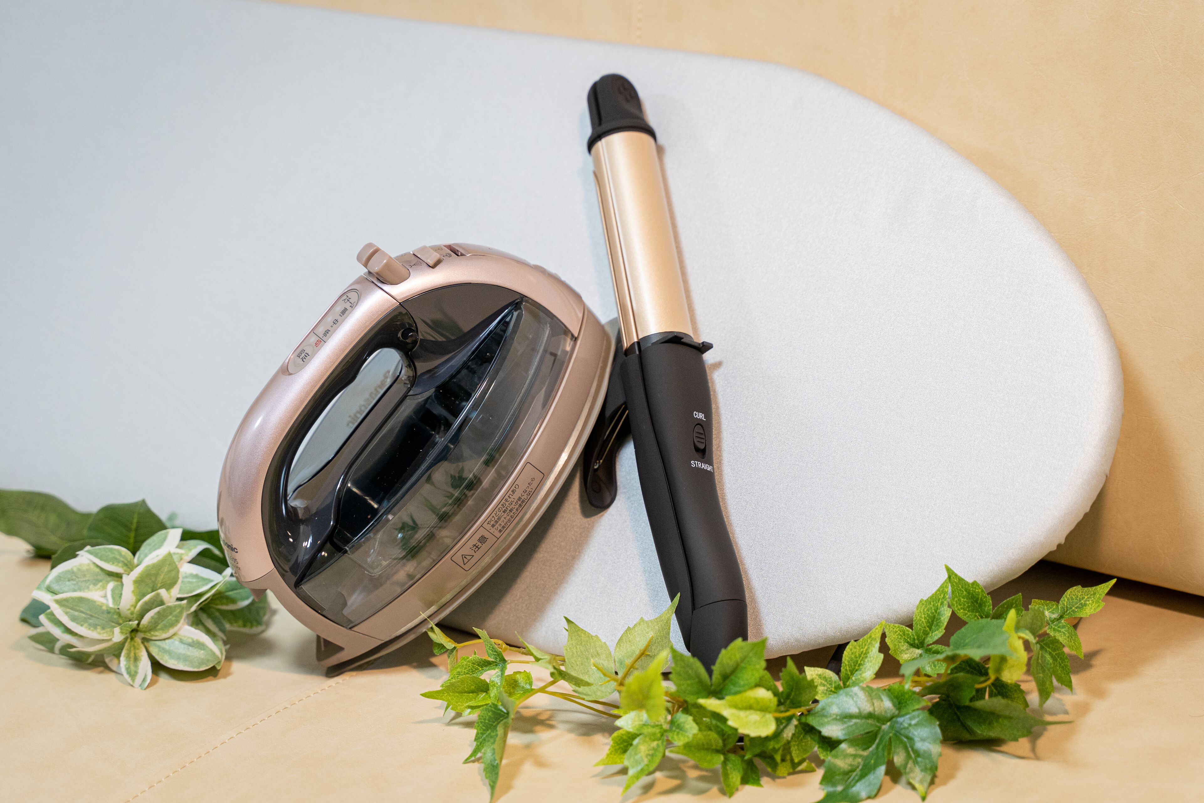 A hair dryer and a curling iron placed on a beige surface with green plants.
