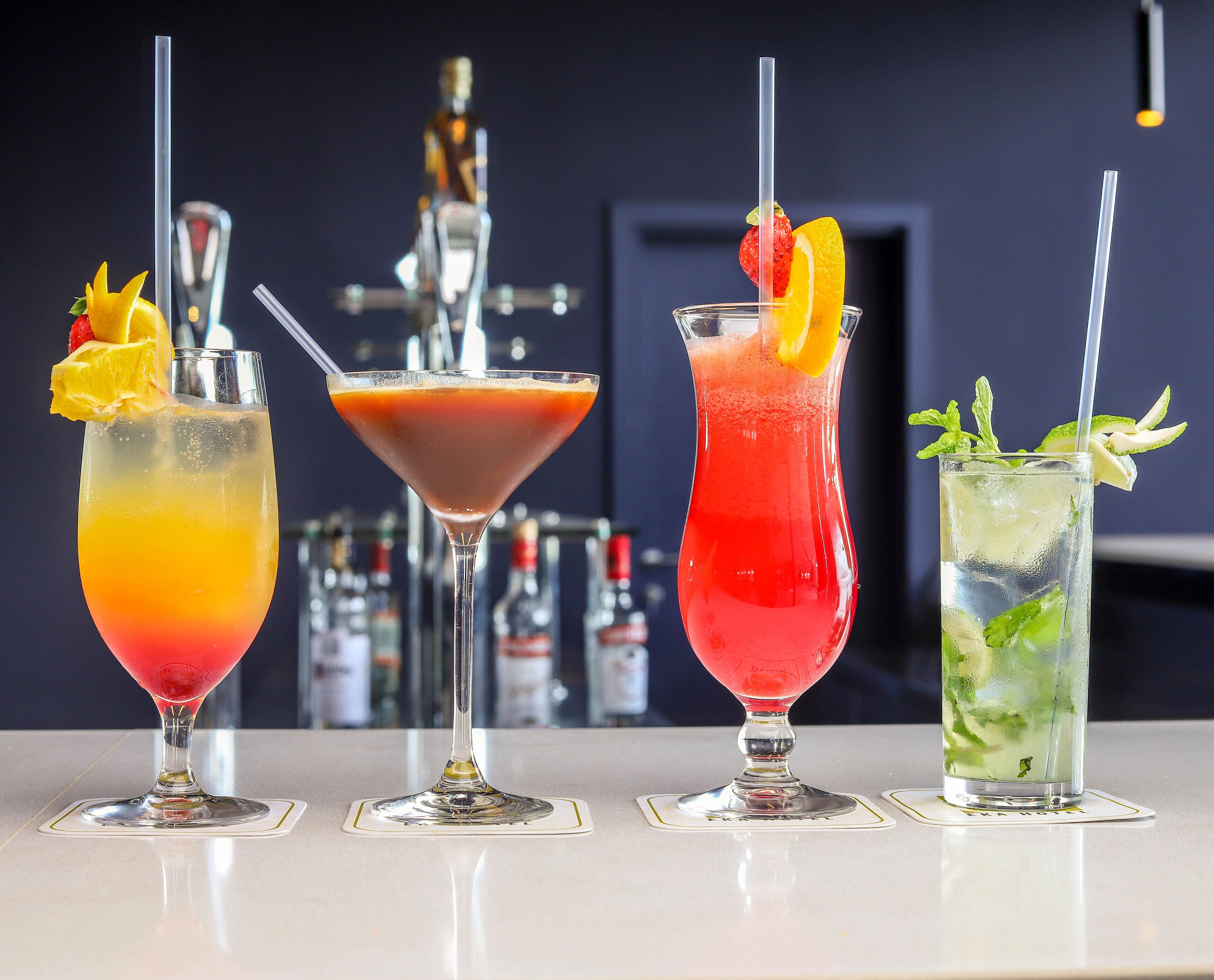 Four cocktails on a bar counter with a blurred background of bar equipment.