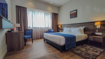 A hotel room with a bed, bedside table, lamp, chair, and a desk with a lamp and flowers.