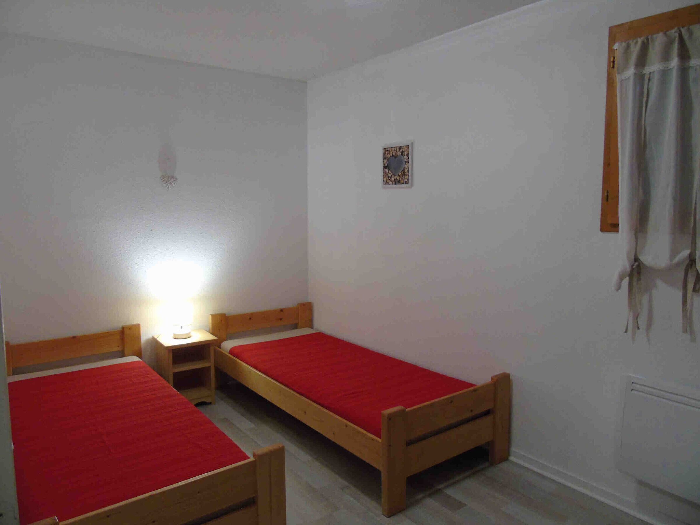A small, simple room with two wooden beds, each with a red bedspread. There is a small table with a lamp, and a window with a curtain.
