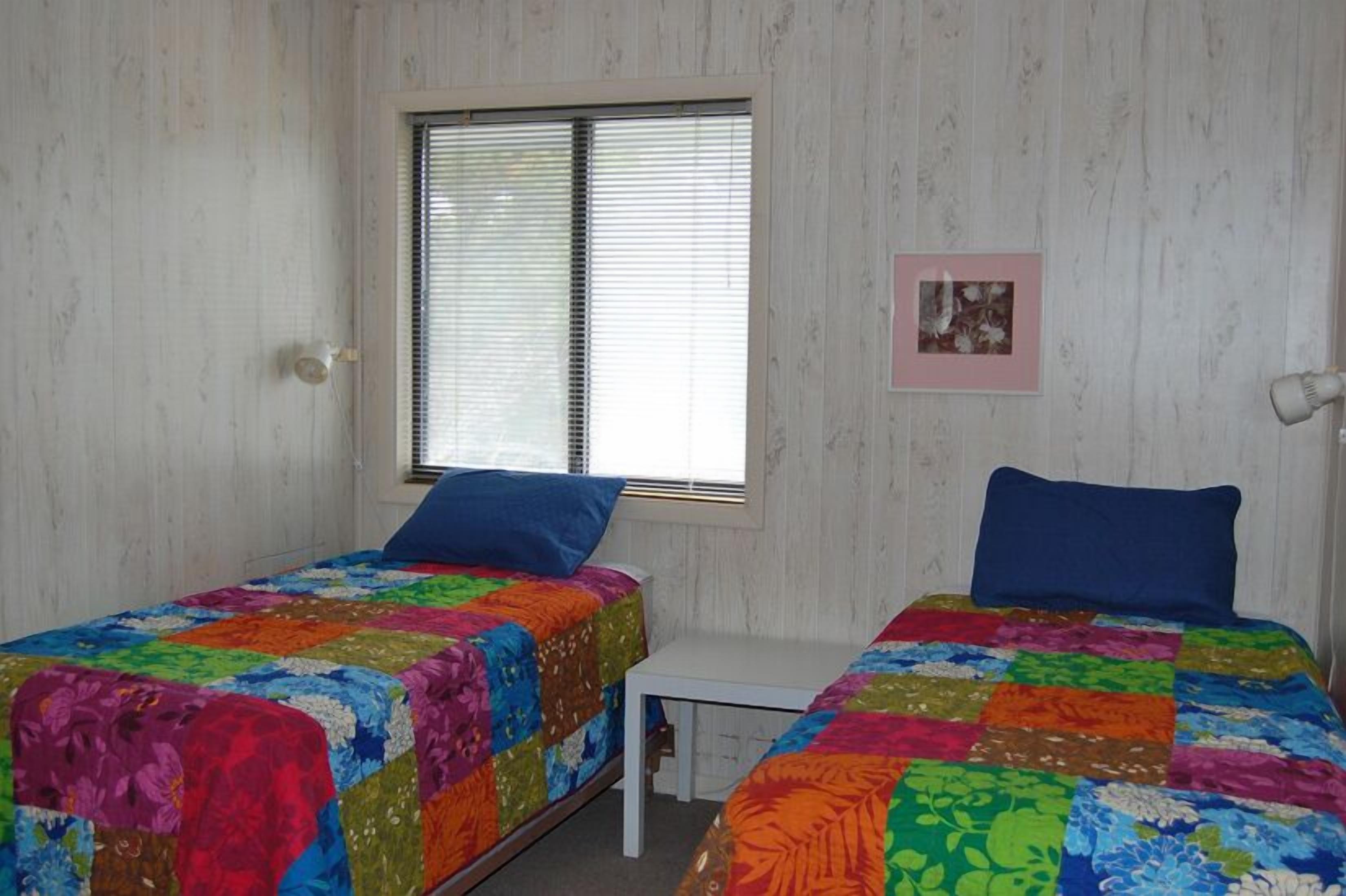 Une chambre avec deux lits, chacun recouvert d’une couverture aux couleurs vives, une petite table blanche, une fenêtre avec des stores et un tableau encadré au mur.