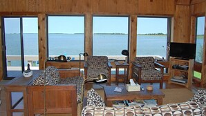 Een houten chalet met grote ramen met uitzicht op een wateroppervlak, ingericht met banken, een salontafel en een televisie.