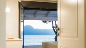 A room with a wooden floor, a bed, a bedside table with a candle, and a view of a lake and mountains through an open door.