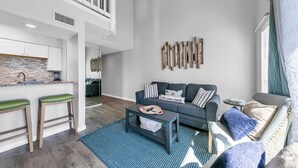 Ein modernes Wohnzimmer mit grauem Sofa, einem Couchtisch und einem blauen Teppich. Es gibt eine Küchenzeile mit weißen Schränken und einer Spritzschutzleiste.