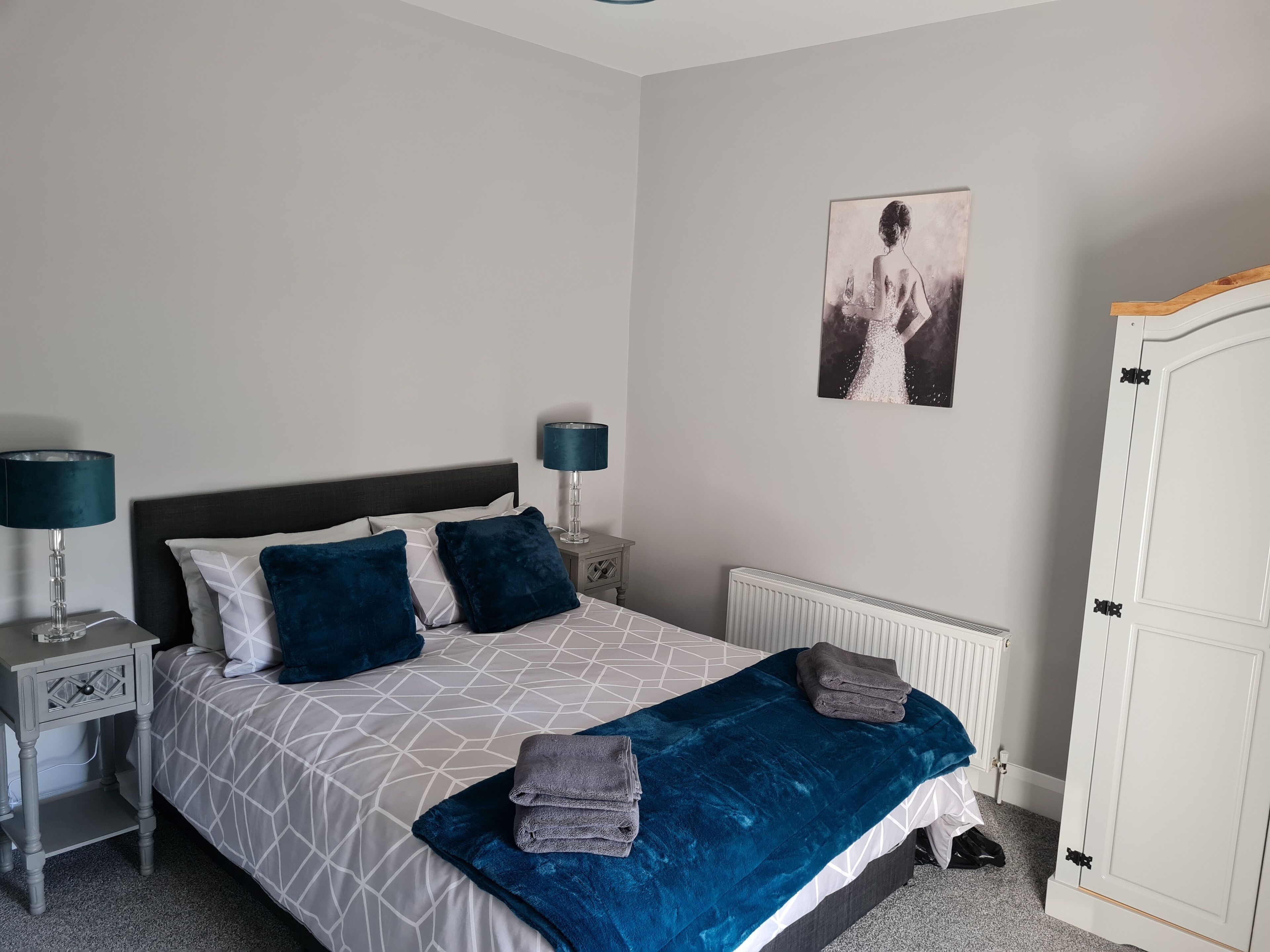 En pænt redt seng med et sengetæppe med geometrisk mønster, to mørkeblå puder og et foldet håndklæde. Der er to sengeborde med lamper, en radiator og en hvid garderobe.