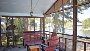Uma área de estar coberta ao ar livre, com deck de madeira, uma cadeira de balanço, um banco e uma mesa.