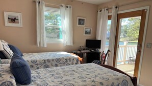 A bedroom with two beds, a desk, a TV, and a view of the outdoors. - Horsin Around | 3BR with Private Dock, Deck and Pet-Friendly (Chincoteague)