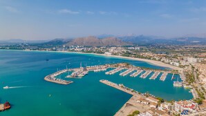Vue aérienne d’une marina côtière avec des bateaux amarrés, une plage de sable et une vue sur la ville en arrière-plan.