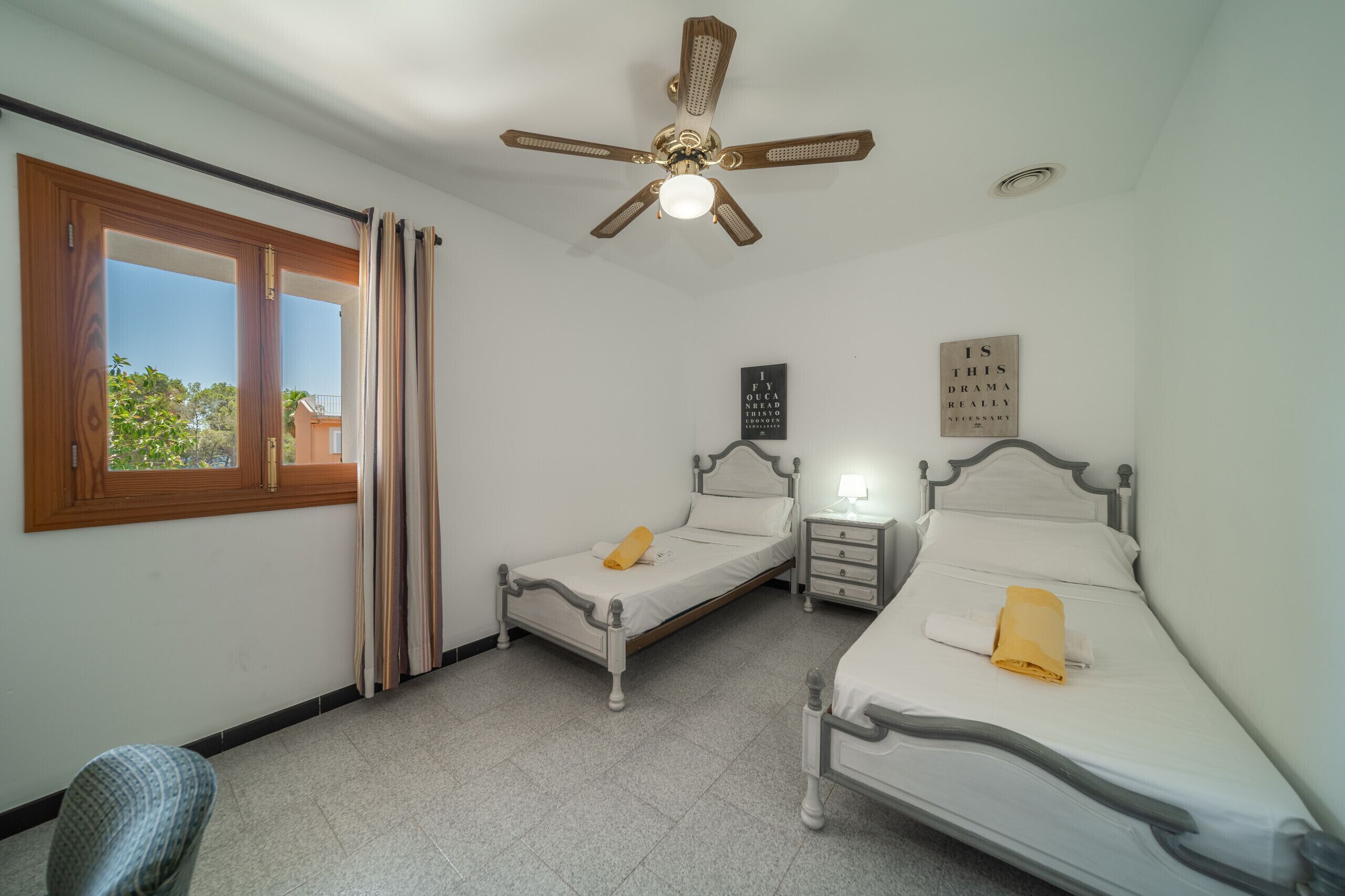 A bedroom with two single beds, a ceiling fan, a nightstand with a lamp, and a window with curtains.