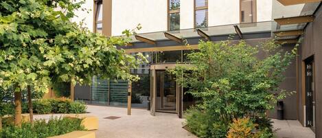 A modern building with a glass entrance, surrounded by greenery and a concrete pathway.