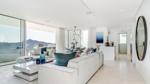 Un salon moderne avec un grand canapé blanc, une table basse en verre et une vue sur le paysage urbain, visible à travers de grandes fenêtres.