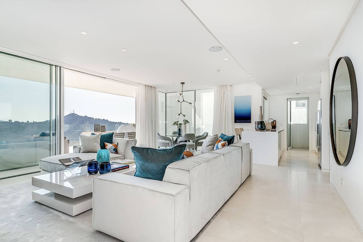 Una sala de estar moderna con un sofá blanco grande, una mesa de centro de vidrio y vistas a un paisaje urbano a través de ventanas amplias.