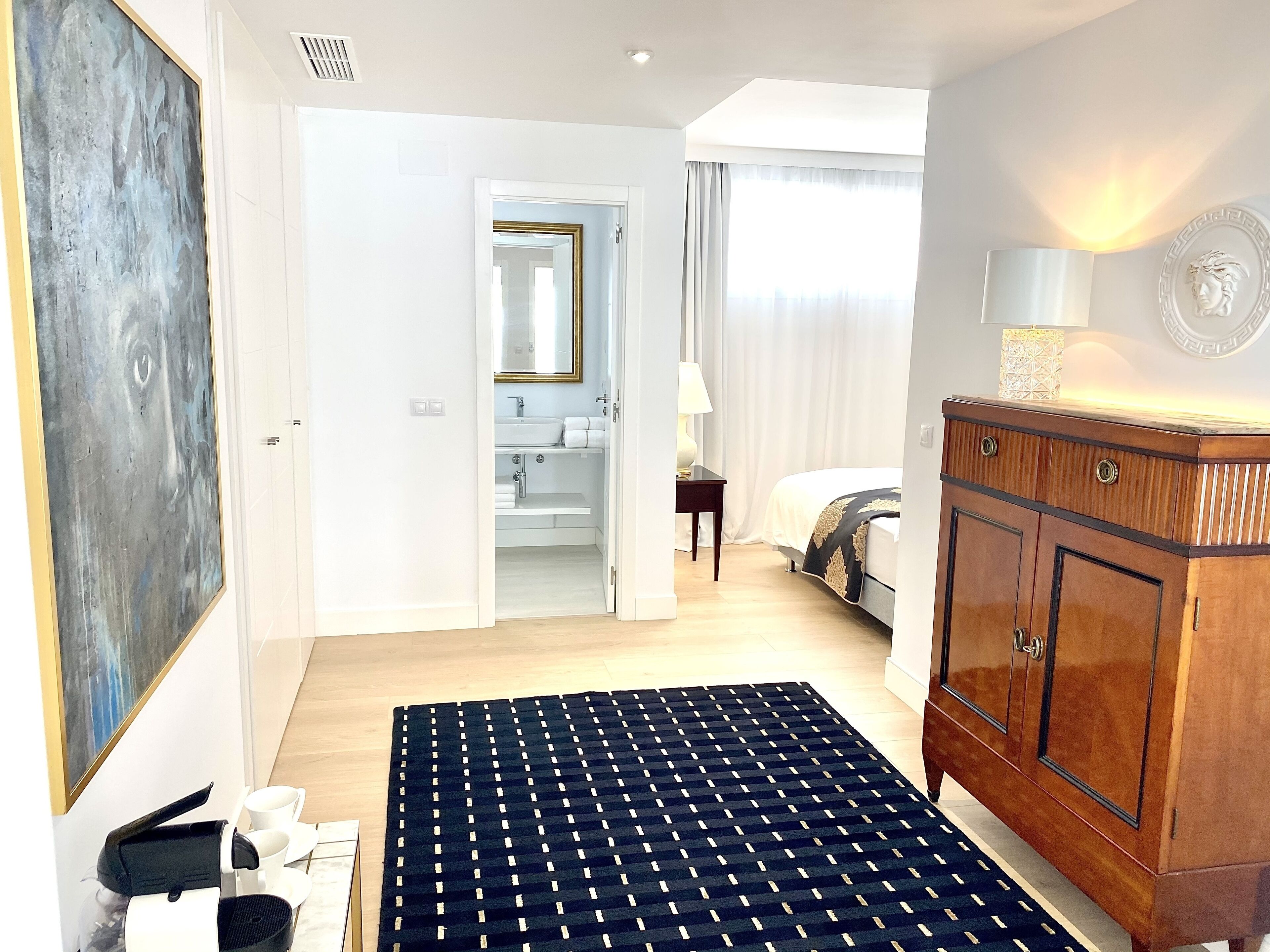 Habitación de hotel con un cuadro abstracto grande, un armario de madera, una cama con edredón estampado, un baño con espejo y lavabo, y vistas al exterior a través de una ventana.