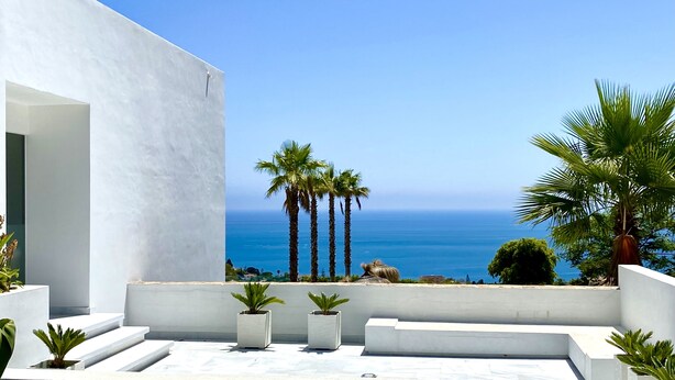 Une terrasse extérieure moderne offrant une vue dégagée sur l’océan, les palmiers et un bâtiment blanc.