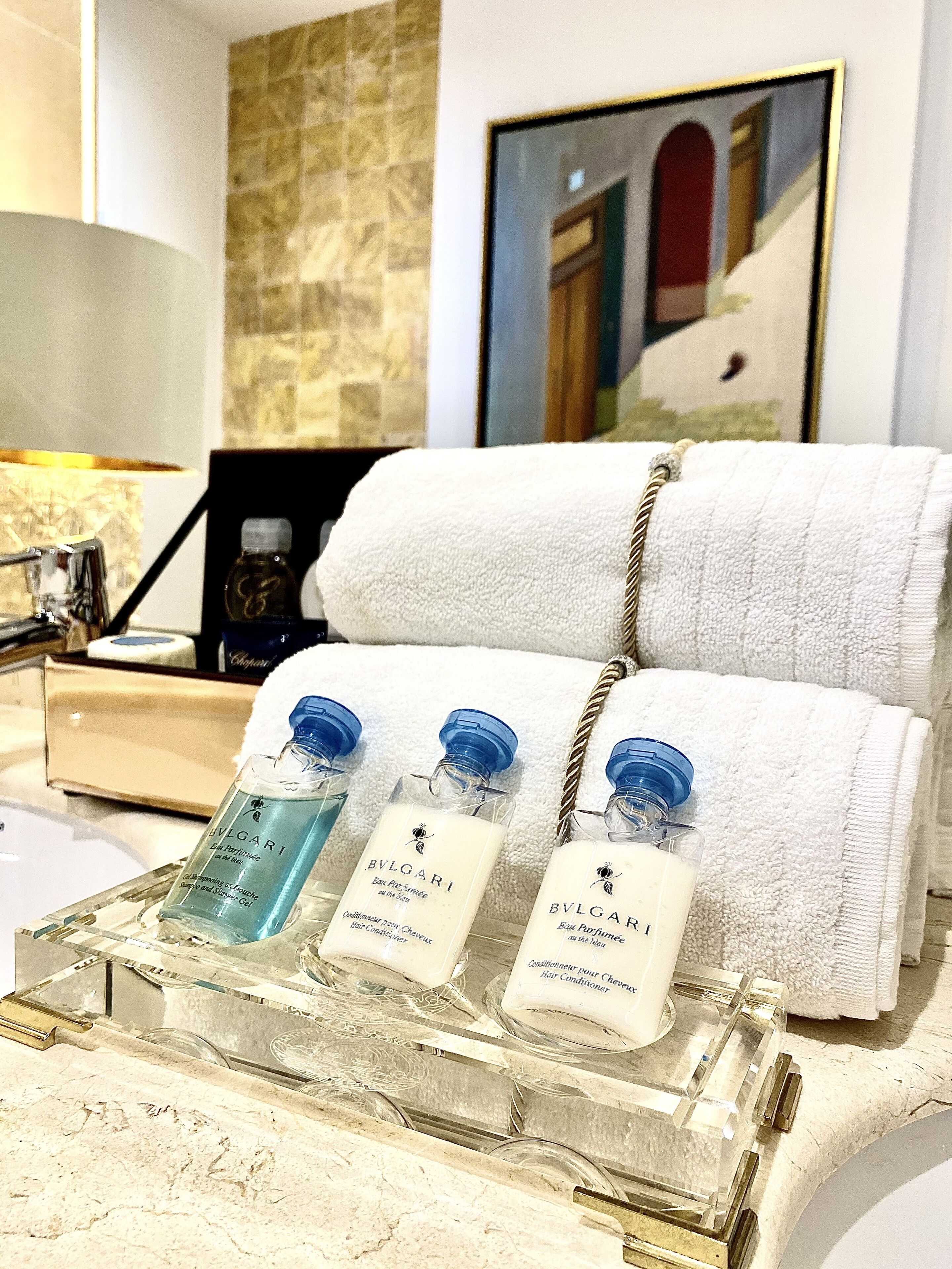 Un baño con encimera de mármol, tres botellas de acondicionador para el cabello Bvlgari, una toalla blanca y un cuadro en la pared.