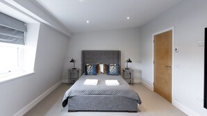 A bedroom with a large bed, two bedside tables, a window with blinds, a wooden door, and a ceiling light.