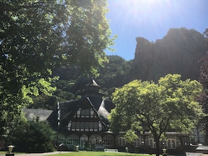 Ein traditionelles Gebäude mit steilem Walmdach, einem Turm und Rundbogenfenstern, umgeben von üppigem Grün und einer markanten Felsformation im Hintergrund.