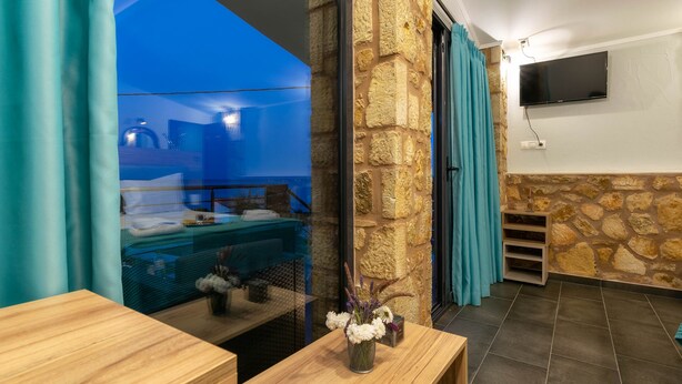 A modern room with a stone wall, a wooden desk, a vase with flowers, a mounted TV, and a view of the outside. - Georgia Apartments (Zakynthos)