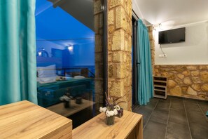 A modern room with a stone wall, a wooden desk, a vase with flowers, a mounted TV, and a view of the outside. - Georgia Apartments (Zakynthos)