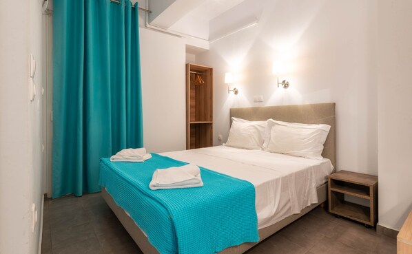 A neatly made bed with a teal blanket and white pillows, a wooden nightstand, and a teal curtain. - Georgia Apartments (Zakynthos)