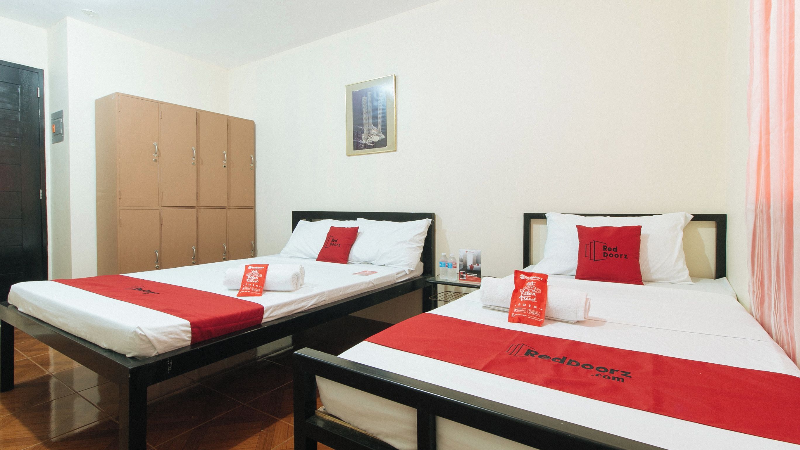 Two single beds with red bedspreads, a wooden wardrobe, and a framed picture on the wall.