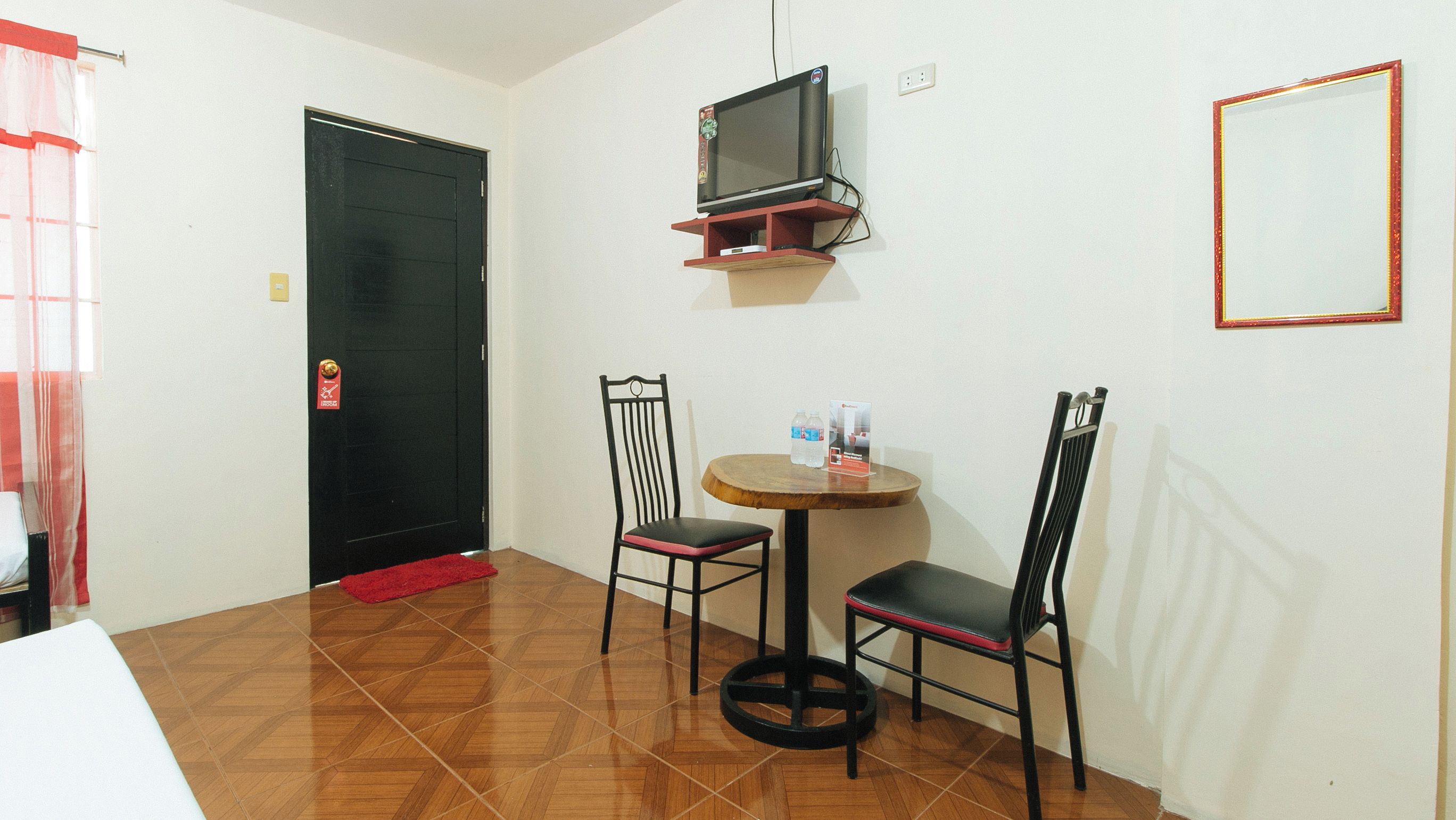 A small room with a black door, a television mounted on the wall, two chairs around a small round table, and a red doormat.
