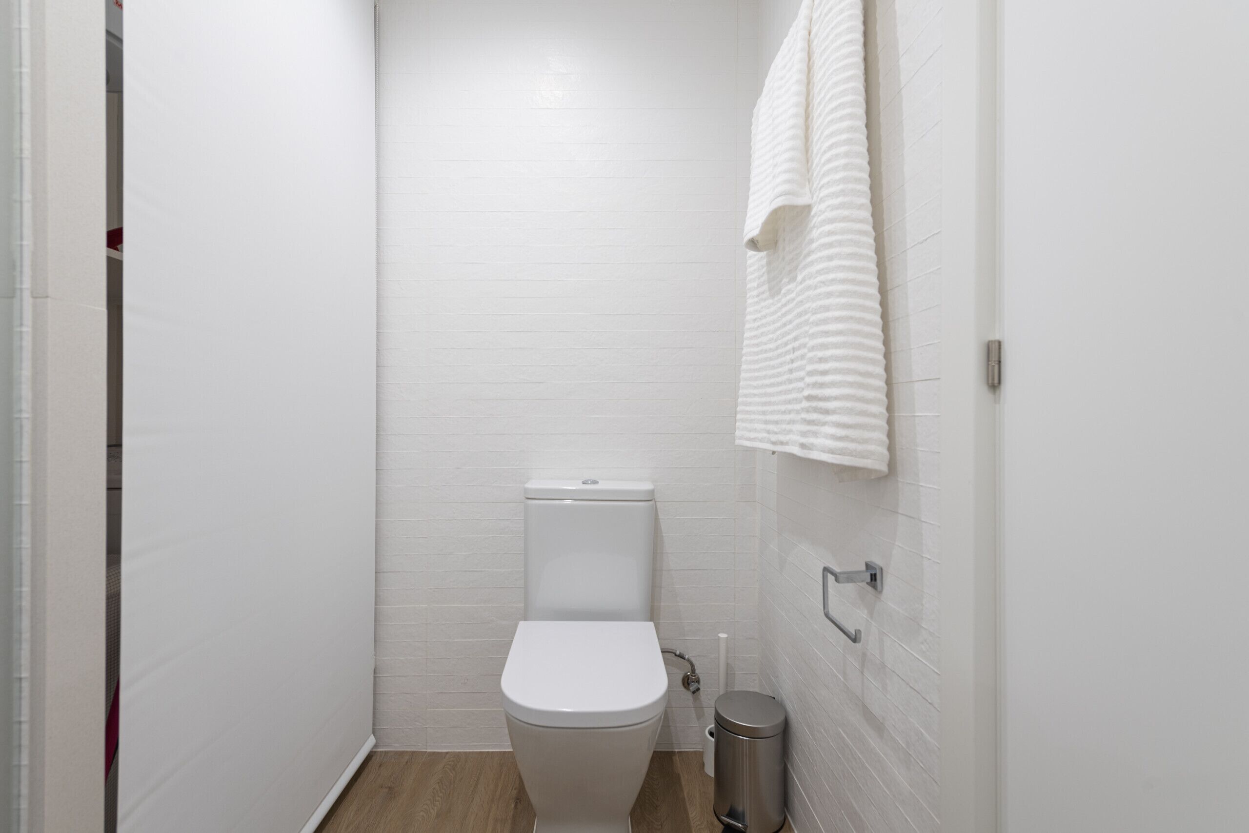 Un baño pequeño con paredes blancas, un inodoro, un cubo de basura y una toalla colgada en la pared.