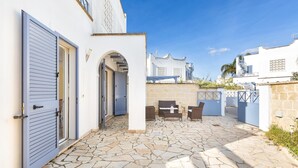 Un patio con suelo de piedra, una zona de estar con mesa y sillas, y un edificio blanco con puerta azul.
