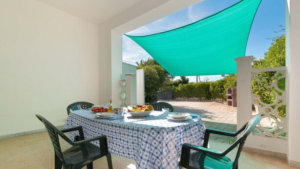 Un coin repas avec une table dressée pour deux personnes, recouverte d’une nappe à carreaux bleus et blancs, et une voile d’ombrage verte au-dessus.
