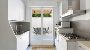 A compact kitchen with white cabinets, a gas stove, a microwave, and a dining area visible through a glass door. - 2150 Casa Mandù (Manduria)