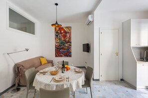Zona pranzo con un tavolo rotondo apparecchiato per quattro persone, un divano con un cuscino giallo, una lampada da terra e un quadro astratto colorato sulla parete.