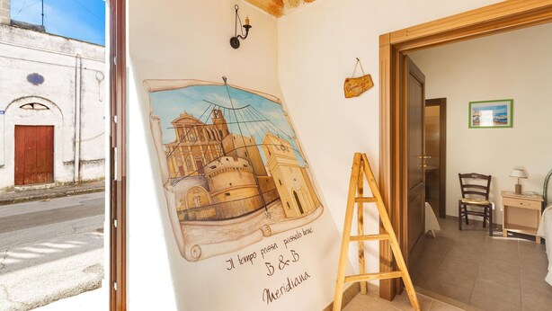 Ein Zimmer mit einer Wandmalerei eines historischen Gebäudes, einer Holzleiter, einem Stuhl und einem kleinen Tisch mit einer Lampe.