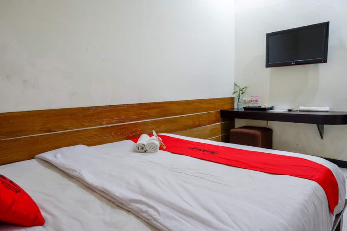A neatly made bed with a red and white bedspread, a red pillow, and a rolled towel. There is a wooden headboard, a flat-screen TV mounted on the wall, and a small desk with a vase and remote controls.